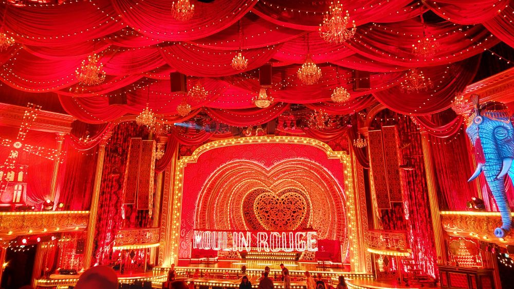 A picture from the inside of the Moulin Rouge musical dome in Cologne. The color scheme is different shades of red and gold. There's a blue elephant on the right side of the room (like the one in the movie), and the Moulin Rouge windmill on the left side. At the back of the room, you can see the stage with a background of hearts and the name of the musical displayed. The ceiling is covered in red drapes and is decorated with golden chandeliers that are hanging above the audience. There's also strings of fairy lights, and the room in general has a lot of lights in a warm golden tone. 
