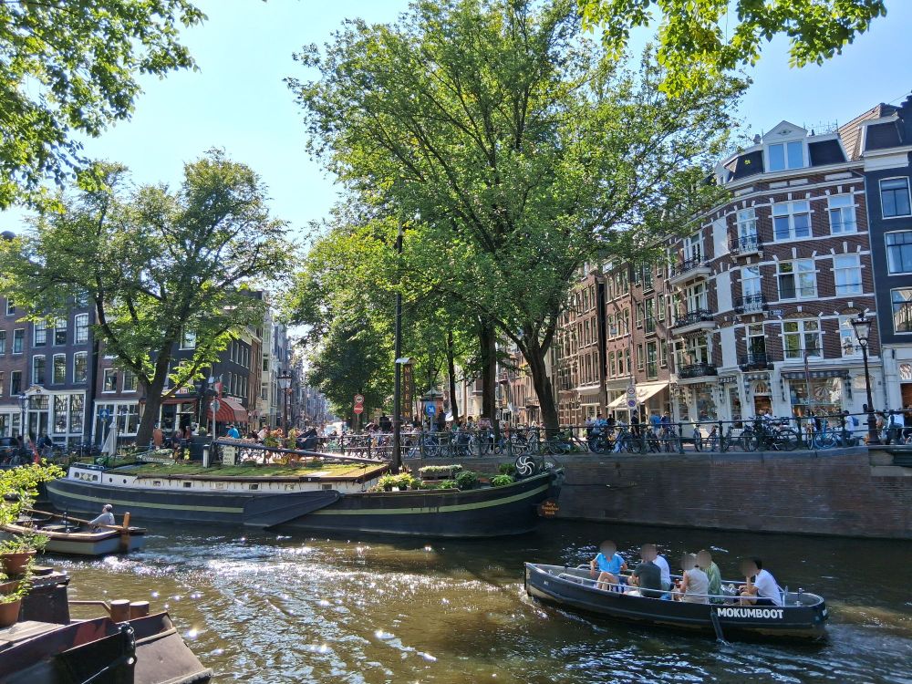 A picture of boats on a canal in Amsterdam (I think this one was the Prinsengracht). There's two few smaller boats with people on them, and a bigger houseboat in the back.