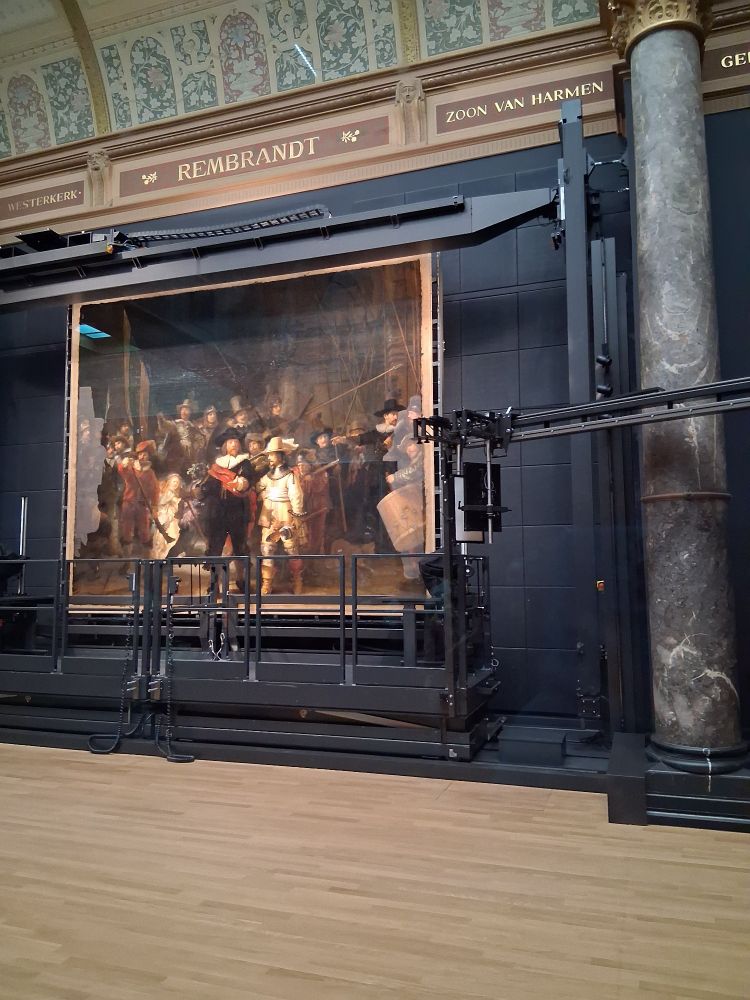 A picture of the painting 'The Night Watch' (1642, oil on canvas) by Rembrandt van Rijn (1606 - 1669). It is described by the museum as follows:

"Rembrandt's largest and most famous painting was made for one of the three headquarters of Amsterdam's civic guard. These groups of civilian soldiers defended the city from attack. Rembrandt was the first to paint all of the figures in a civic guard piece in action. The captain, dressed in black, gives the order to march out. The guardsmen are getting into formation. Rembrandt used the light to focus on particular details, like the captain's gesturing hand and the young girl in the foreground. She was the company mascot. The nickname Night Watch originated much later, when the painting was thought to represent a nocturnal scene." 

The painting is currently behind glass walls, because conservators are working on it. There are five phases total to renew the painting. At the moment, it's in phase 3: removal of varnish and overpaint.

"In the current phase of Operation Night Watch, conservators are removing the old varnish layers. Varnish is a transparent layer that protects and saturates the paint layers so the colours are perceived in their correct brightness and intensity."

You can see some gray areas in the painting where the varnish has already been removed.