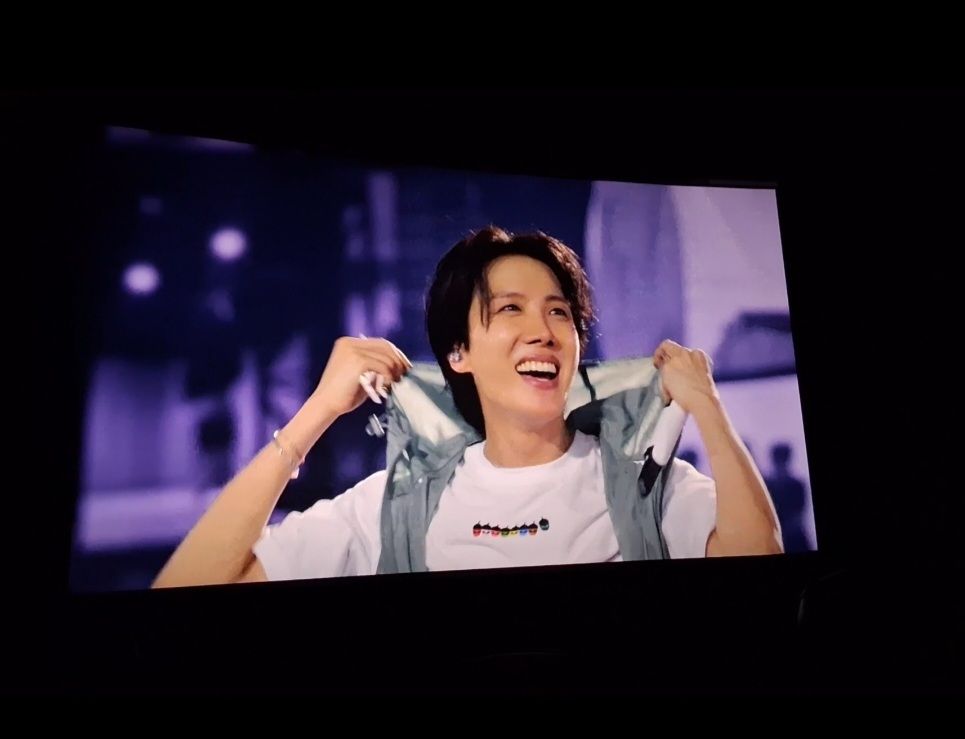 A picture I took at the movie theater with j-hope on the screen. It shows a moment from the encore of his Hope on the Stage concert in Seoul. He's wearing his own tour merch, a white t-shirt with the tour stop acorns on it, and a gray wind vest with a hood. He's grabbing the vest's hood and is smiling brigthly at army in his sunshine style. 