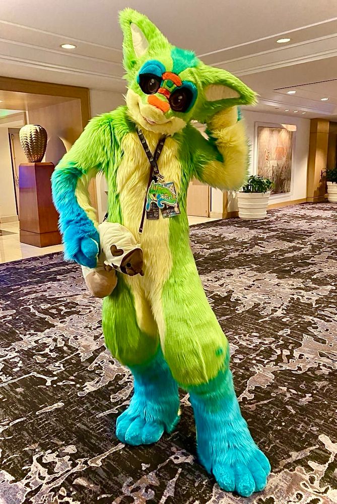 An eared day gecko stands in a hotel hallway scratching his head with a confused look while a large plush grub hangs by his waist. 