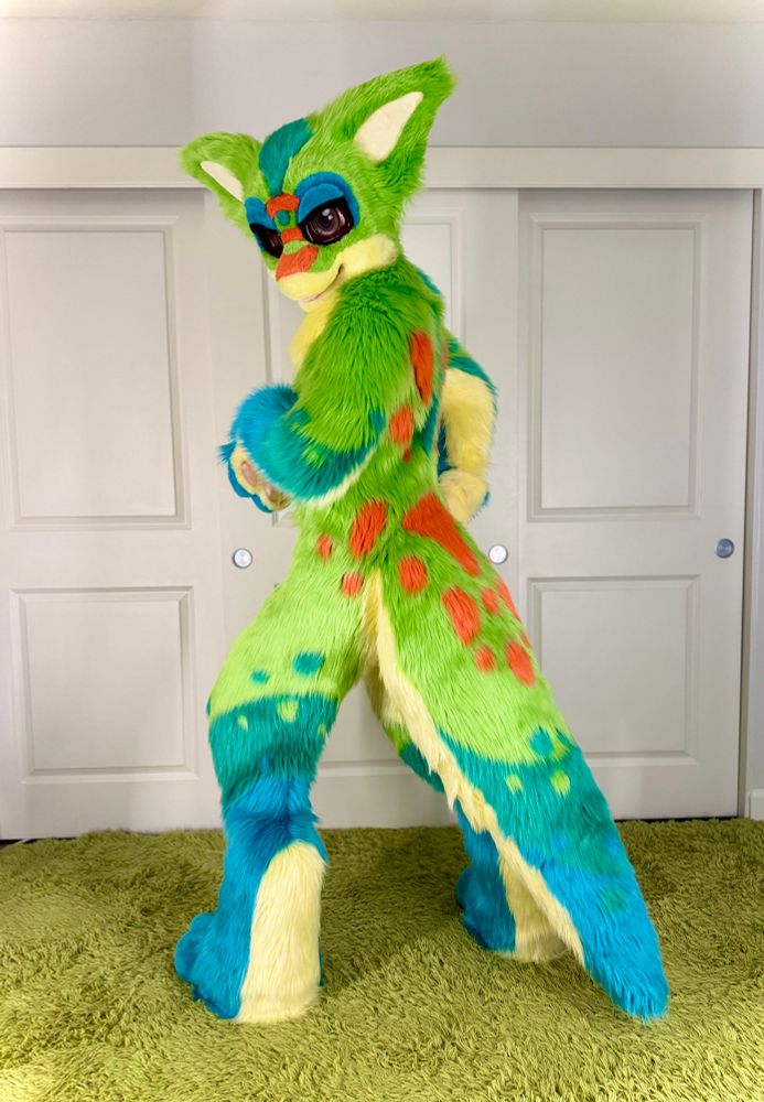 Basyl, a big eared, anthro day gecko stands atop greenery with his back facing the camera, showing off his lovely orange, green, and blue spotted pattern and big tail. 