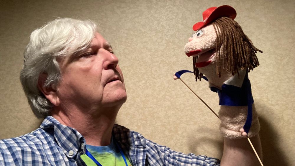 A gray haired man considers a sock puppet flawlessly outfitted as Agent Carter.