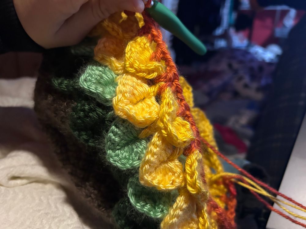 A hand holding a half finished crochet project and a crochet hook. The project has a brown base and so far has dark and light green as well as yellow leaves, and orange stitches on top. 