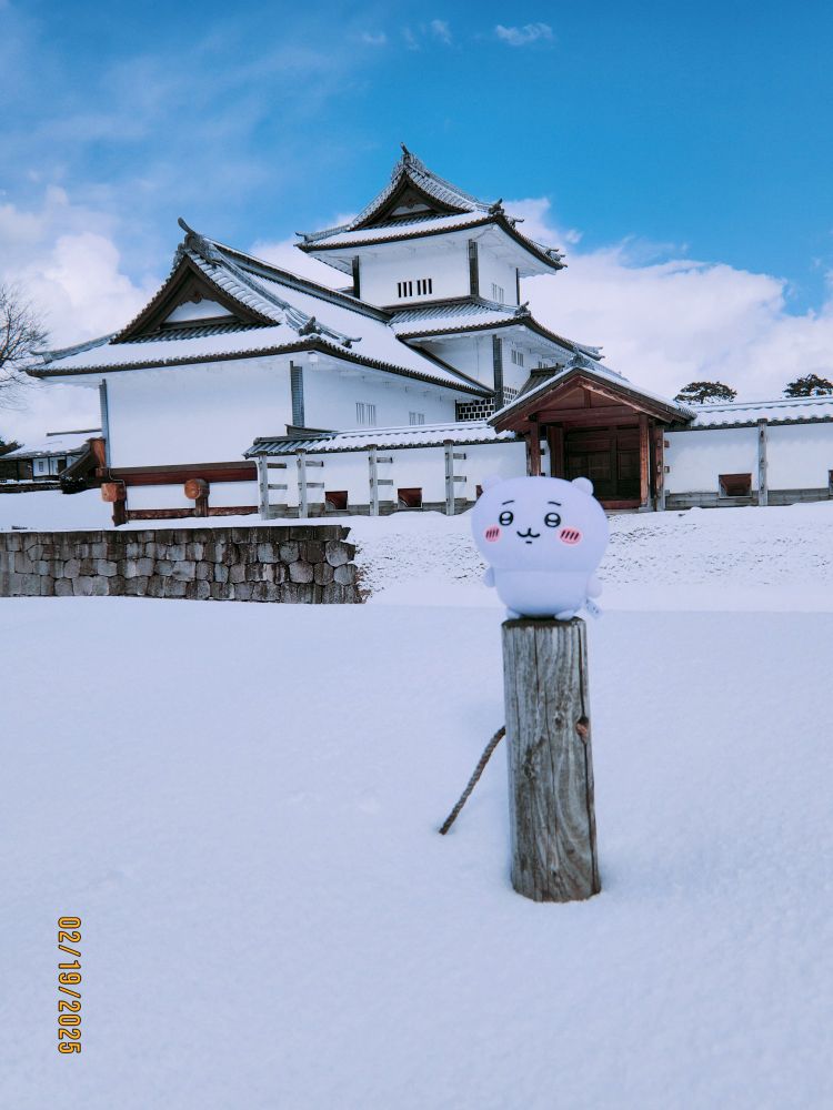 Chiikawa, a small, round plushie with a cheerful expression and rosy cheeks, sits atop a wooden post surrounded by a thick layer of snow. In the background, the majestic Hashizume-mon Gate of Kanazawa Castle stands with its traditional white walls, dark wooden accents, and sloping tiled roofs. The snowy landscape, combined with the blue sky and scattered clouds, creates a serene winter scene, highlighting the contrast between the castle's historical architecture and Chiikawa's playful presence.