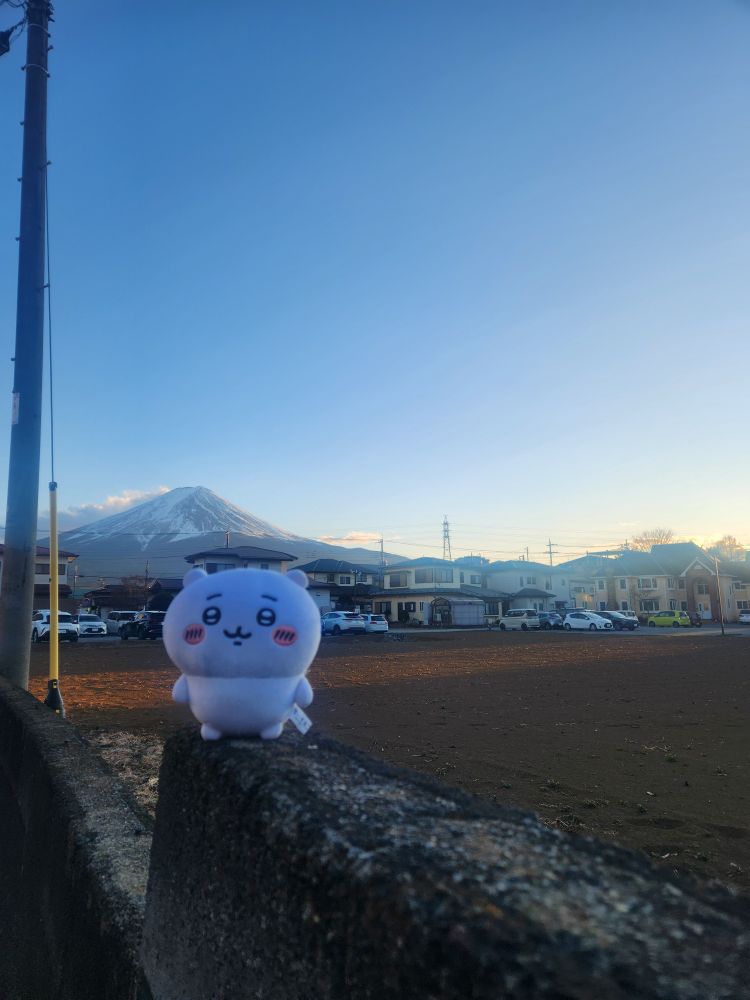 A soft, round plushie named Chiikawa sits on a textured concrete ledge in the foreground. Chiikawa has a cheerful expression with rosy cheeks. Behind it, a clear blue sky stretches above a row of suburban houses and parked cars. In the background, the iconic snow-capped peak of Mount Fuji rises, bathed in the warm hues of the setting sun, creating a serene and picturesque scene.