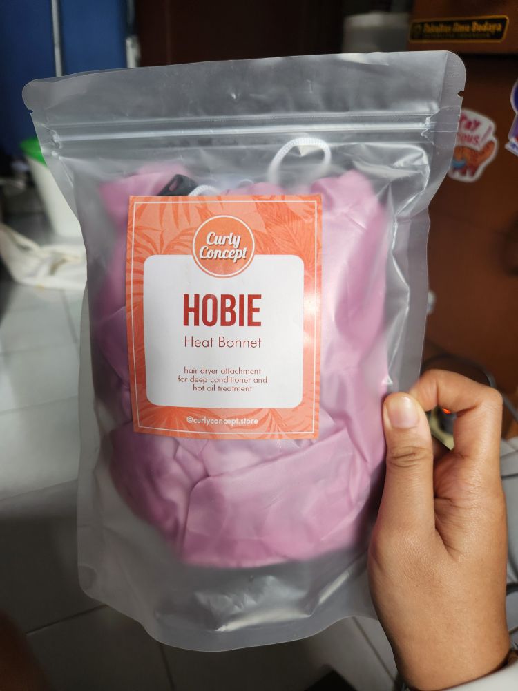 A picture of a hand holding a clear plastic pouch containing a pink heat bonnet. The label on the pouch reads "Curly Concept. Hobie Heat Bonnet. Hair dryer attachment for deep conditioner and hot oil treatment," with the store Instagram handle @curlyconcept.store at the bottom.