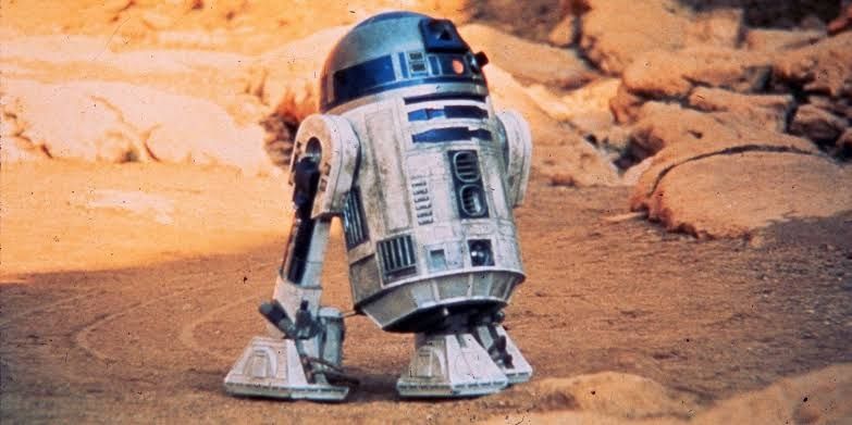 R2-D2, personagem icônico de Star Wars, em um ambiente desértico.

A imagem mostra um robô de formato cilíndrico com pernas curtas. Sua cabeça é arredondada e eu corpo é branco e azul, com diversos botões e luzes piscantes. O robô está em um terreno árido e rochoso, com marcas de suas pegadas na areia. O fundo da imagem é um pouco mais escuro, com rochas e areia de coloração avermelhada.