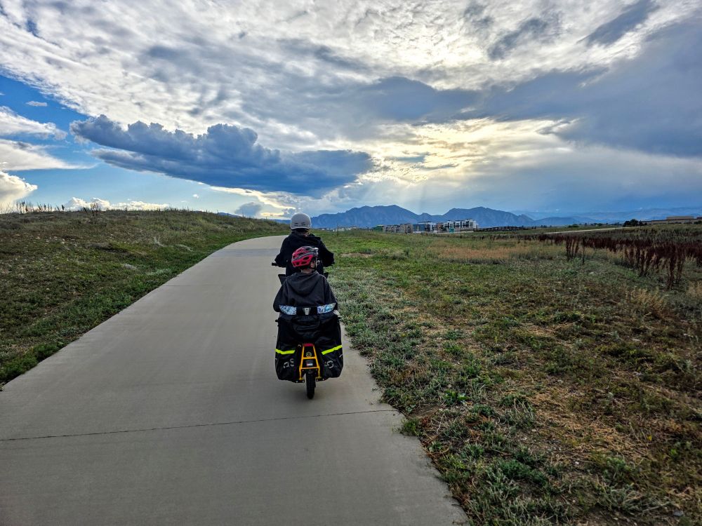 A picture of a cargo bike heading away from the camera on a large bike path going west toward the mountains in the background with a setting sun and lots of dark and bright rain clouds. Towards Boulder, Colorado
