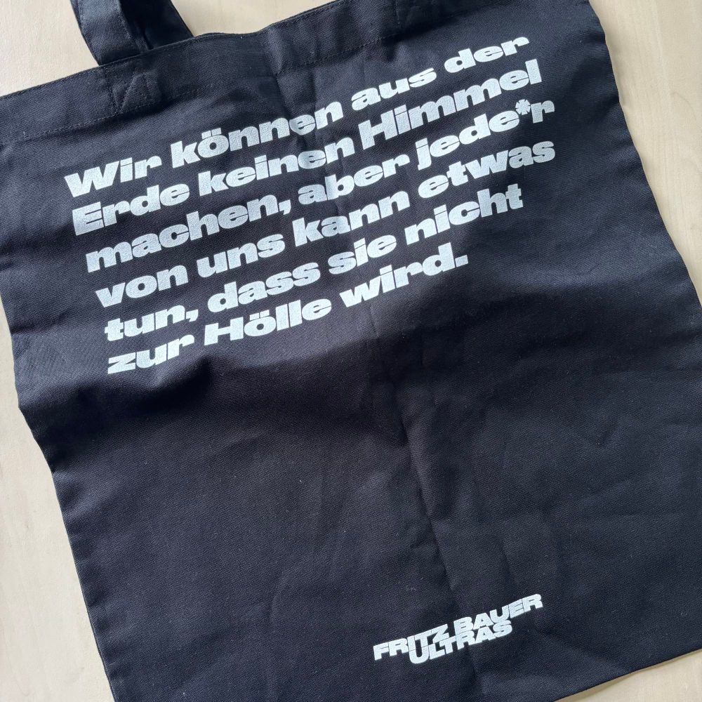 Es handelt sich um ein Foto eines schwarzen Jutebeutels mit dem Aufdruck: Wir können aus der Erde keinen Himmel machen, aber jede*r von uns kann etwas tun, dass sie nicht zur Hölle wird. // Fritz Bauer Ultras