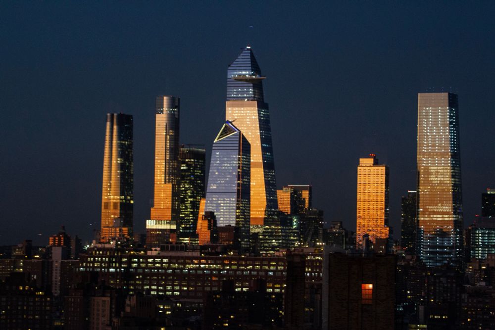 A photograph of New York City at dusk.  The tall skyscrapers reflect the deep orange glow of sunset.  You can see the hundreds of lights in each building 
