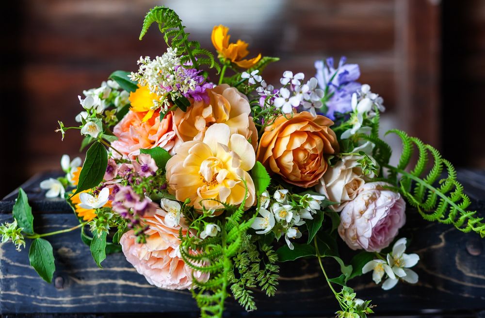 A horizontal shallow focus image of a garden bouquet hanging over a distressed black wood surface, with a rustic wood wall in the background. The bouquet is full of roses of various colors and forms, accompanied by deep orange coreopsis, dame's rockett, vervascum, a large open-centered blue scabiosia, young ferns, mock orange, and privet blooms.
