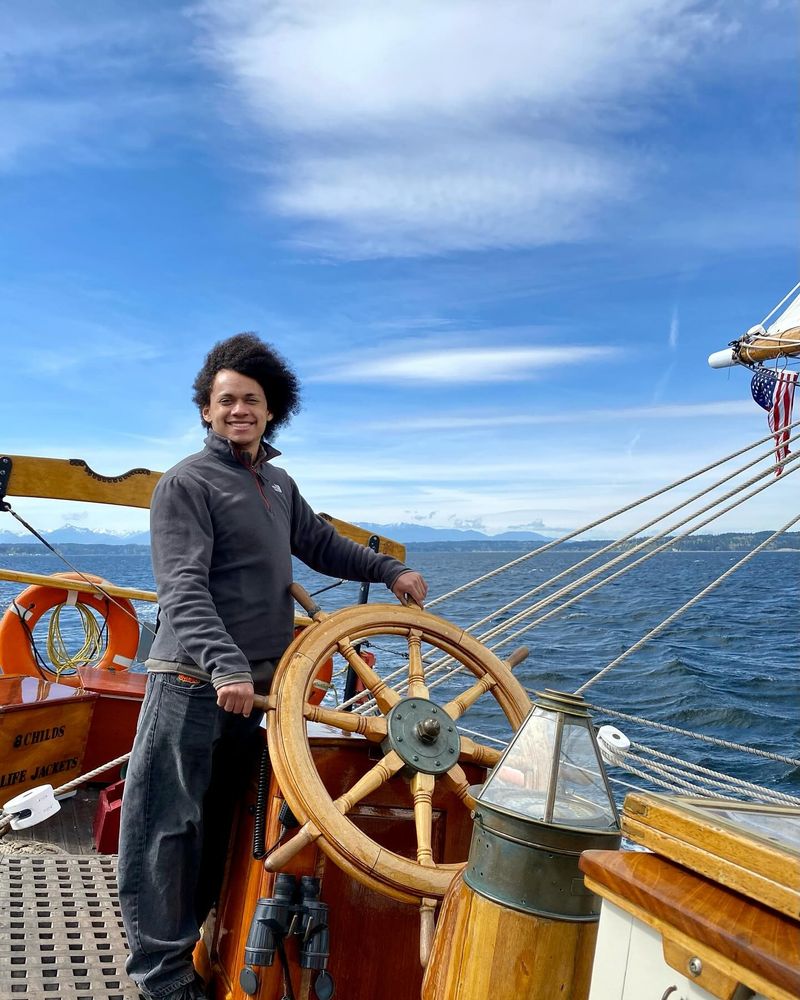 A young man takes the helm of Adventuress under the guidance of her captain and crew. He is wearing layers of warm, black clothing and a very big smile. Adventuress is the platform for education for young folks where they learn about marine science in the Salish Sound region and are introduced to pathways to maritime vocations.
