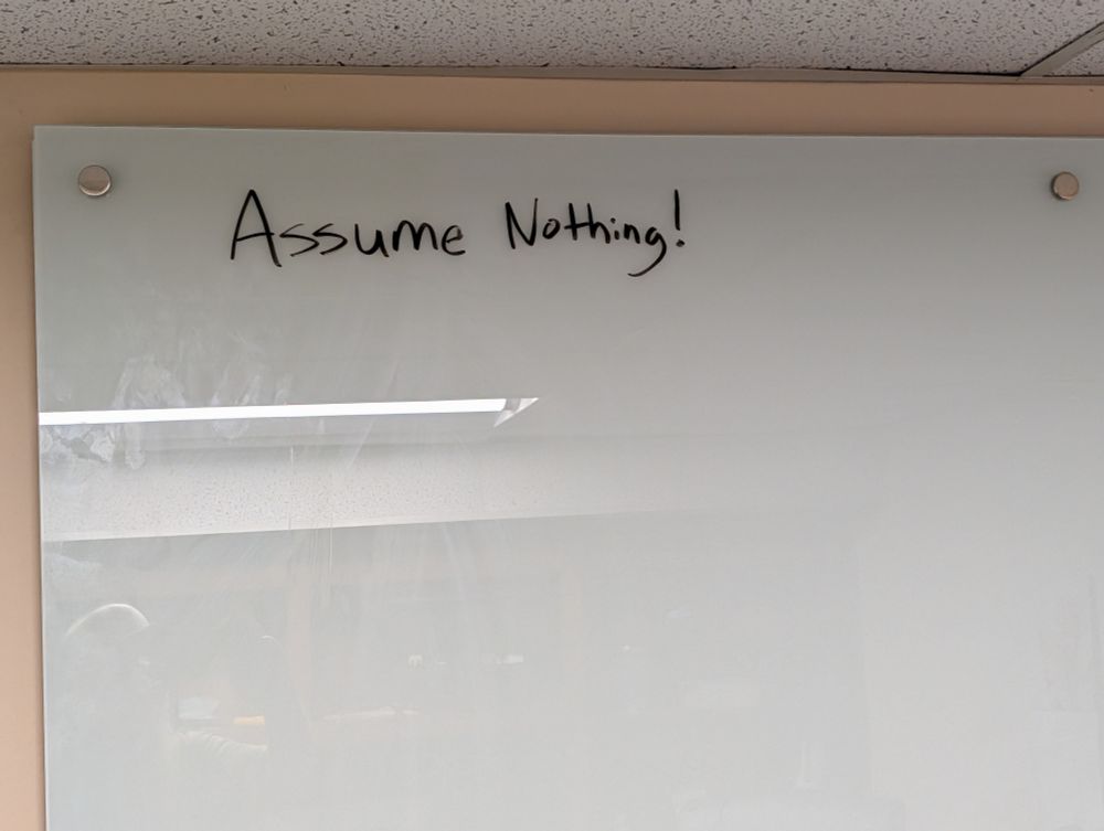 A view of part of a white board where someone else has written "assume nothing!" in black dry erase marker