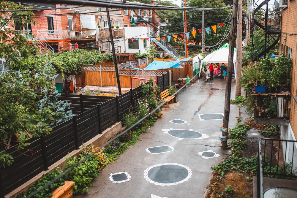 A “green alley” in Montreal. The alley seems to have conventional pavement, but it is lush with greenery, decorated with paint and pennant banners, and appears to be hosting a block party.