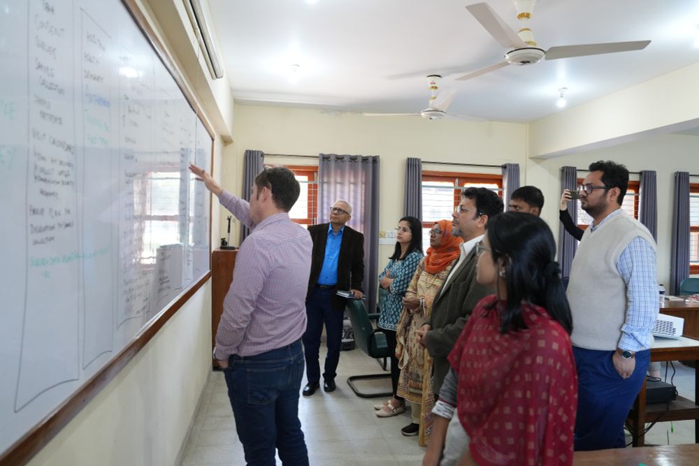 A group of researchers stand in front of a whiteboard. One man is pointing at something on the board and the other seven people are looking on, interested.