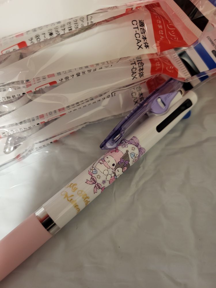 A uni Jetstream three color ink pen with a pastel pink grip and a purple grip is sitting on a package. On the barrel are the Sanrio characters MyMelody and Kuromi. Stacked next to the pen are 4 refills for Tombow Air Touch correction tape dispensers.
