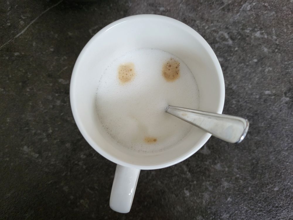 Ein Kaffee im Becher. Von oben fotografiert. Oben zwei Punkte im Milchschaum als Augen, unten ein Punkt als Mund. Als Nase dient der Löffel.