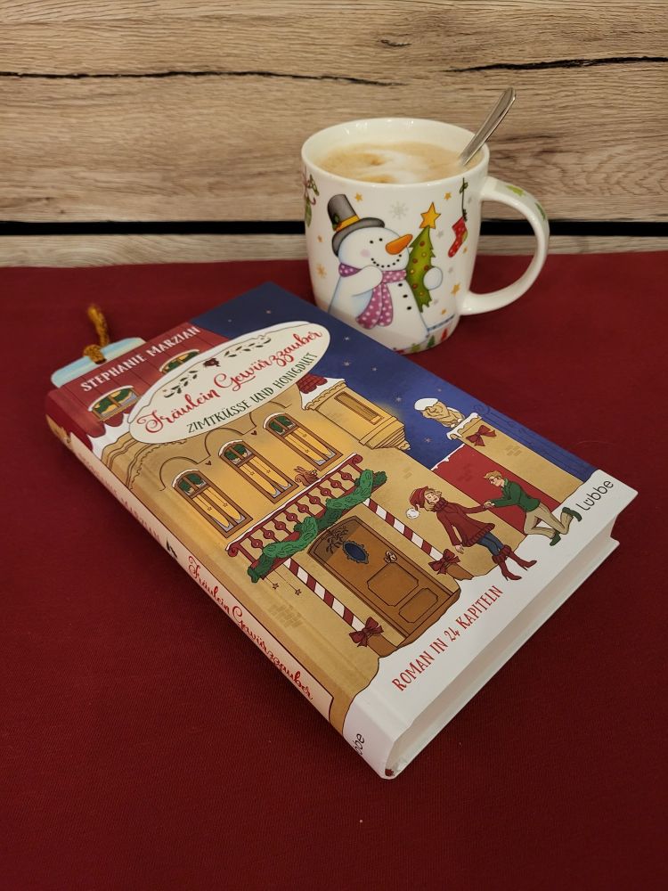 Eine Kaffeetasse, gefüllt mit Cappuccino, mit Schneemandruck darauf. Davor liegt das Buch "Fräulein Gewürzzauber - Zimtküsse und Honigduft" von Stephanie Marzian.