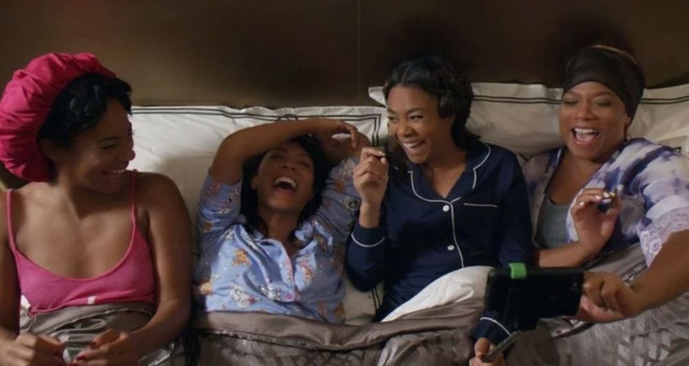 Screenshot from film Girls Trip; 4 beautiful Black women sitting next to each other in a large bed; all are wearing pjs and smiling and laughing