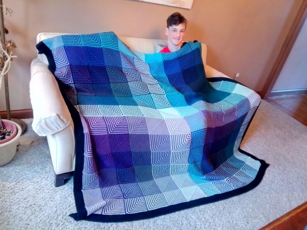 Knitted Hueshift cotton blanket in blues, greens, purples and grays with a black border covering the smiling teen boy it was made for.
