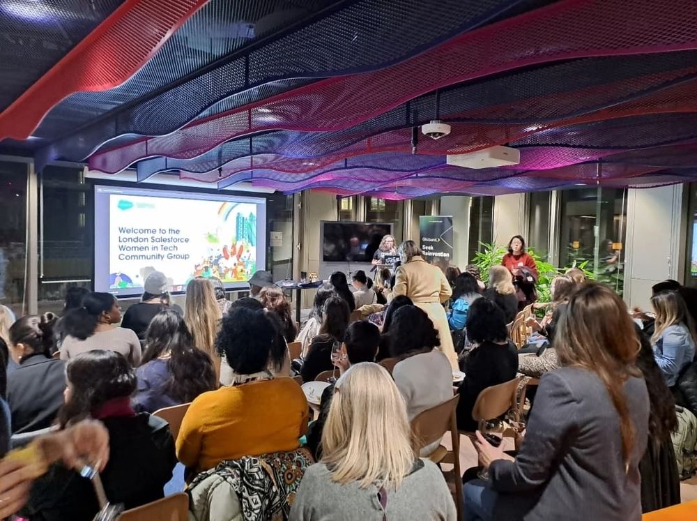 Looking upon on the backs of a group of women sitting watching a lady (Louise) present in front of a big projector screen.