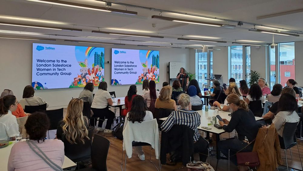 Photo of amazing women sitting in front of Louise Lockie who is standing at the front of a large room, with two big screens on the wall behind her.