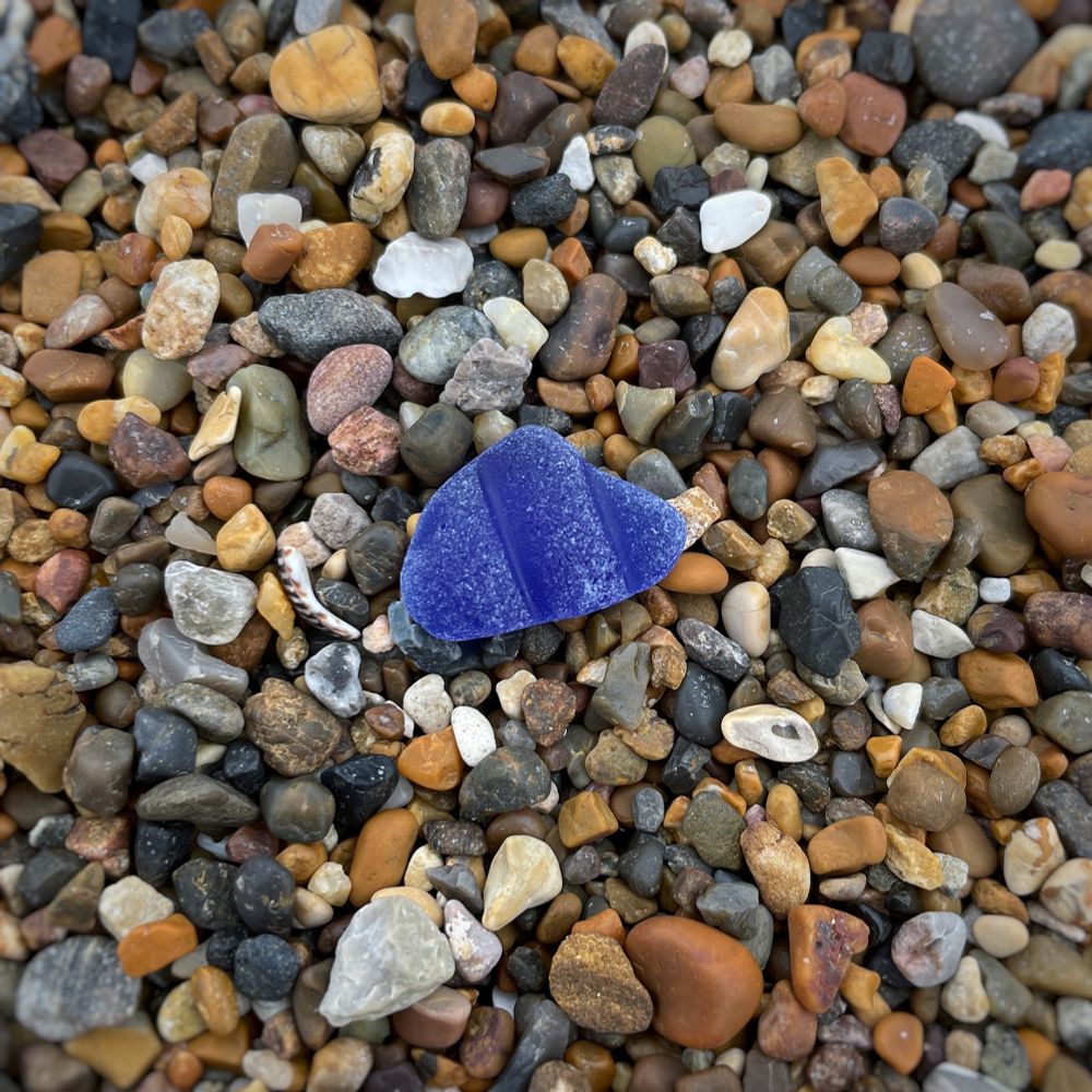 An irregular piece of well frosted primary blue seaglass with smooth fat ridges. It rests in fine gravel in shades of brown, beige and grey