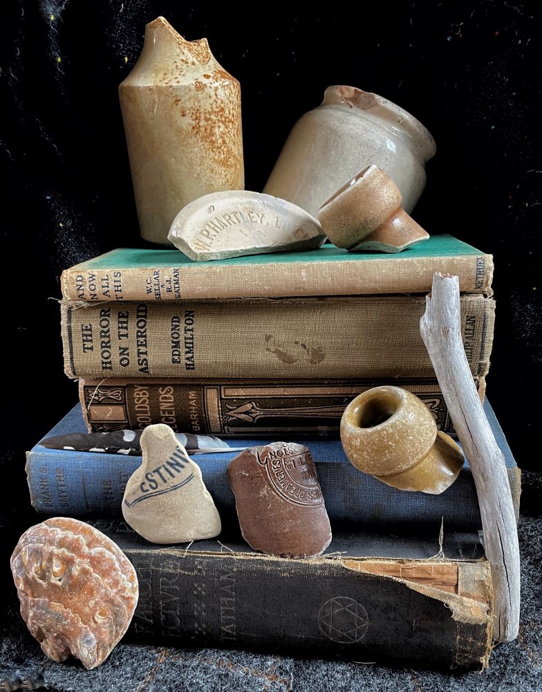 A stack of 5 vintage and antique books (visible titles include “ingoldsby legends” and “the horror on the asteroid”. Balanced on books are partial vintage earthenware jars, large stoneware flagon spouts, parts of things like ceramic marmalade jars. A wand like driftwood stick and pinky-orange oysters-shell also feature 