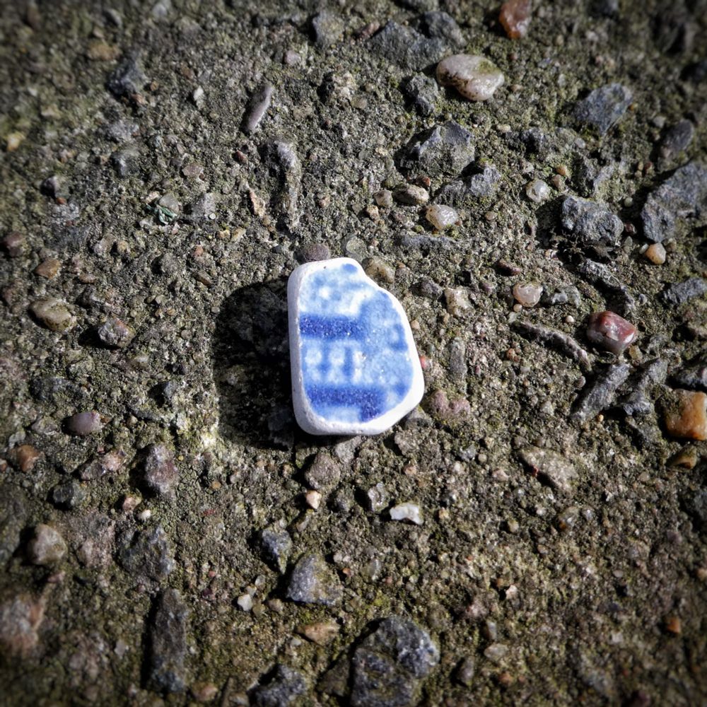 A roughly rectangular piece of sea worn pottery, a blue pattern on white, maybe the edge pattern of willow, maybe something else. It lies in a dark brown-grey rock with pebble inclusions.