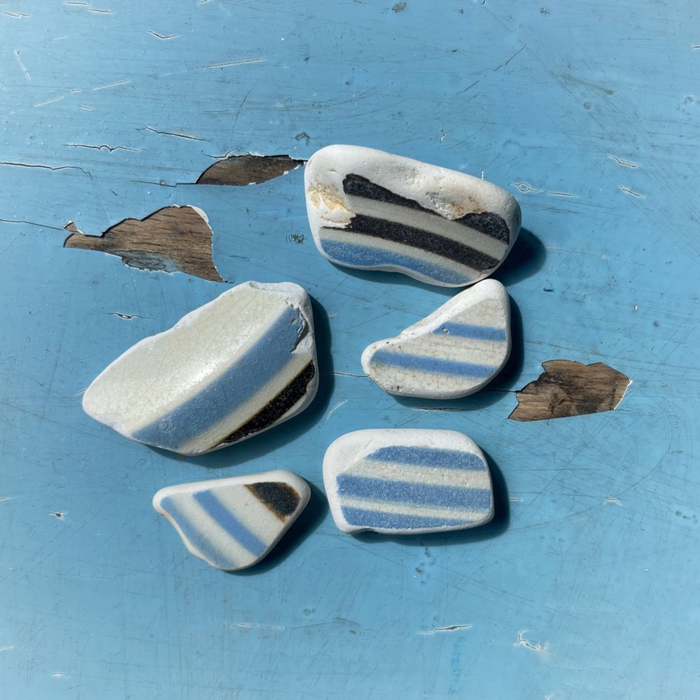 Five pieces of irregularly shaped sea pottery, white with blue and some dark brown stripes. They are laid in a blue painted wooden surface with some paint peeling away