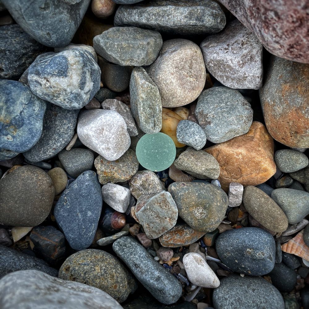A seaworn and frosted aqua glass marble lying among mixed stones and pebbles