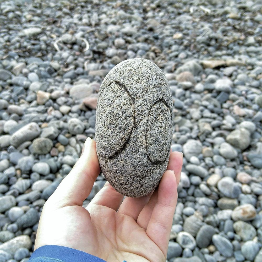 Hand holding an oval granite pebbles with two round dark natural marks looking like a pair of eyes. Background is a very pebbly beach