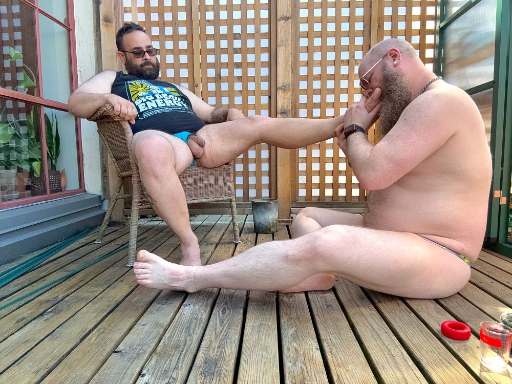Two men, one is seated and the other one is sitting on the deck worshipping his friends feet. 
