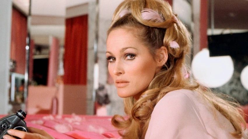 A photo of Ursula Andress as Vesper Lynd (and also James Bond) in 1967's Casino Royalle. She's laying in bed in a nightgown, holding a camera, with curlers in her hair.