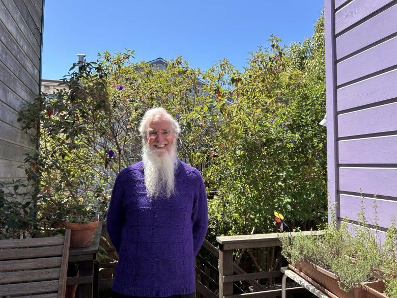 Photo of smiling man with long white beard wearing purple sweater, standing on deck of purple house with purple flowers shrubbery behind.