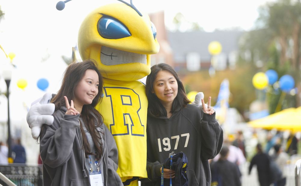 two females holding up the peace sign with the University of Rochester's mascot, Rocky, in between them smiling for a photo.