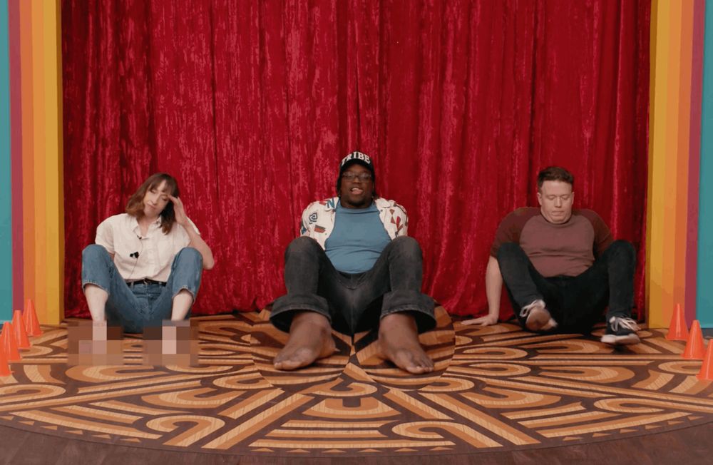 Katie Marovitch, Ify Nwadiwe, and Brennan Lee Mulligan line up at a starting line to crab walk. Katie's bare feet are blurred, Ify's bare feet are magnified, and Brennan is wearing shoes.
