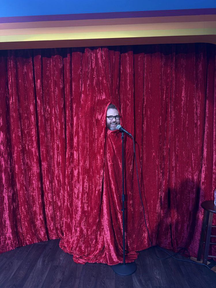 Sam Reich with curtains draped around him so only his head is visible, stands at a microphone.