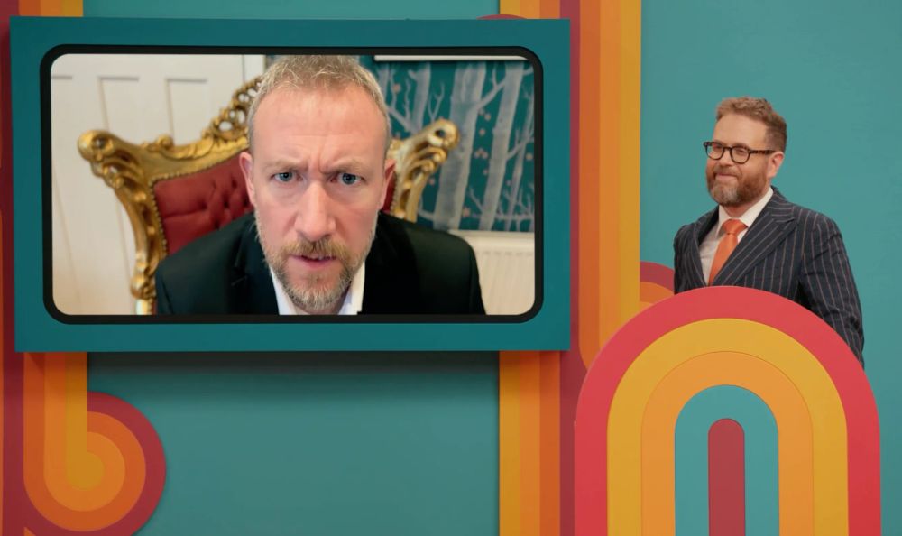 A screenshot of Alex Horne from Taskmaster on the prompt screen from Game Changer, as Sam Reich stands by smiling.