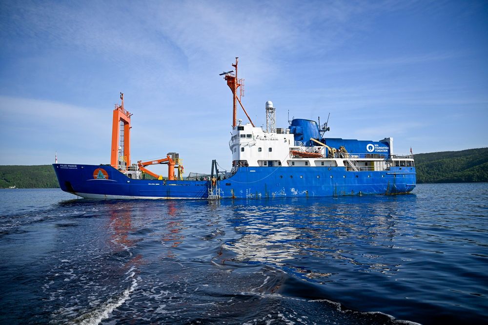 Blue and white research vessel named Polar Prince sailing on calm water with forested hills in the background under a clear sky.