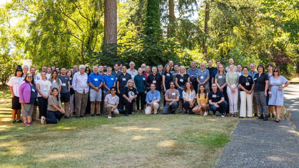 Large group of people posing outdoors on grass with trees and sunlight in the background during daytime event.