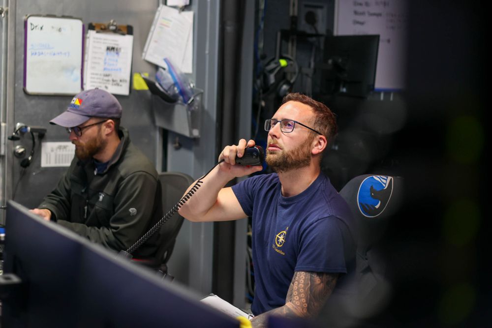 Two people inside a control room; one speaks into a radio while the other works at a computer.