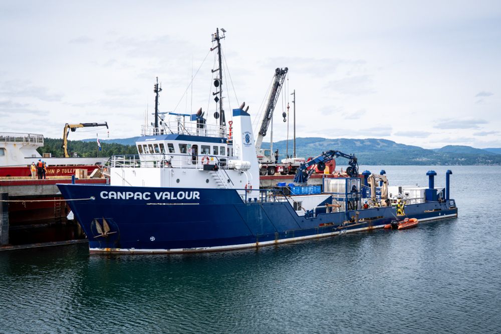 Blue and white vessel named CANPAC VALOUR docked at a harbour with cranes and equipment on board against a mountainous backdrop.
