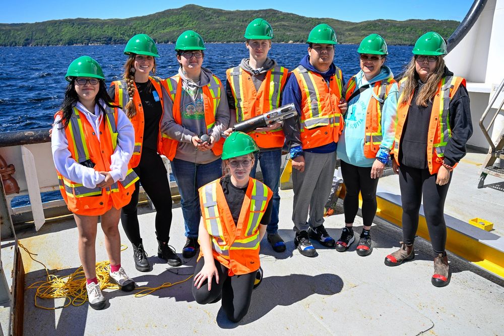 Group of eight people wearing green helmets and orange safety vests standing on a boat deck with water and hills in the background.