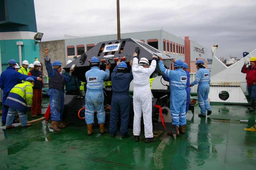 Team of workers in coveralls and helmets positioning a large subsea structure on a ship deck.