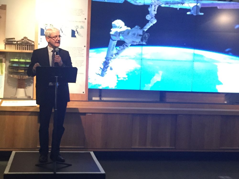 A man in a suit speaking into a microphone next to a screen displaying Canada's space arm.
