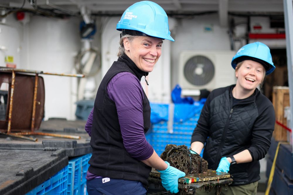 Two smiling people in hard hats and gloves carry a tray with an organic sample aboard a ship.