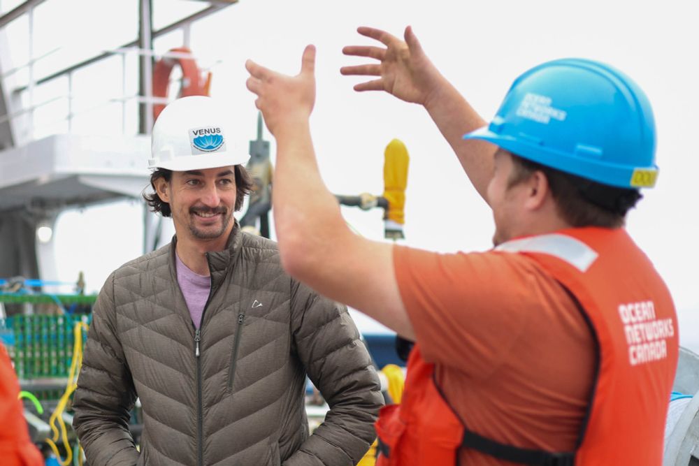 Two people in hard hats and safety gear talk on a ship deck; one gestures animatedly while the other smiles.