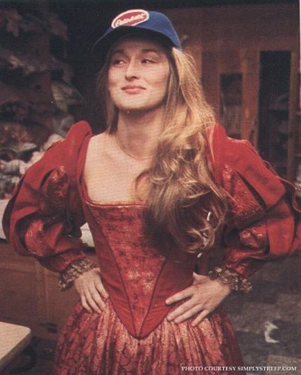 Meryl Streep behind-the-scenes photo from Shakespeare in the Park performance of The Taming of the Shrew. She is wearing a rust(?) colored dress with puffy sleeves and a low neckline, her hair over one shoulder, and a blue baseball cap with the Peterbilt logo on the front.